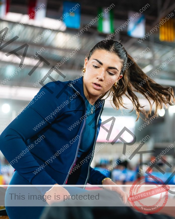 Manuela Blanco Track and field athlete Manuela Blanco sits on a bench indoors, blue jacket and ponytail visible, flags above, blurred track behind.