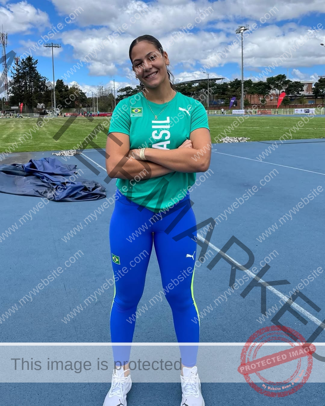 maria-eduarda-dantas-brazil_dudinha_23-1646 Maria Eduarda Dantas, discus thrower from Brazil, stands on a blue track in a green Brazil shirt and blue leggings, arms crossed, smiling.
