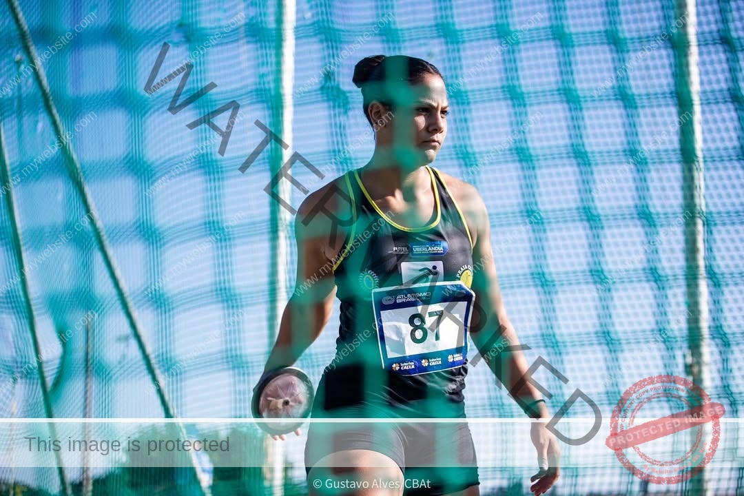 maria-eduarda-dantas-brazil_dudinha_23-1651 Maria Eduarda Dantas, discus thrower from Brazil, stands focused in a netted cage in dark uniform, holding a discus at an outdoor event.