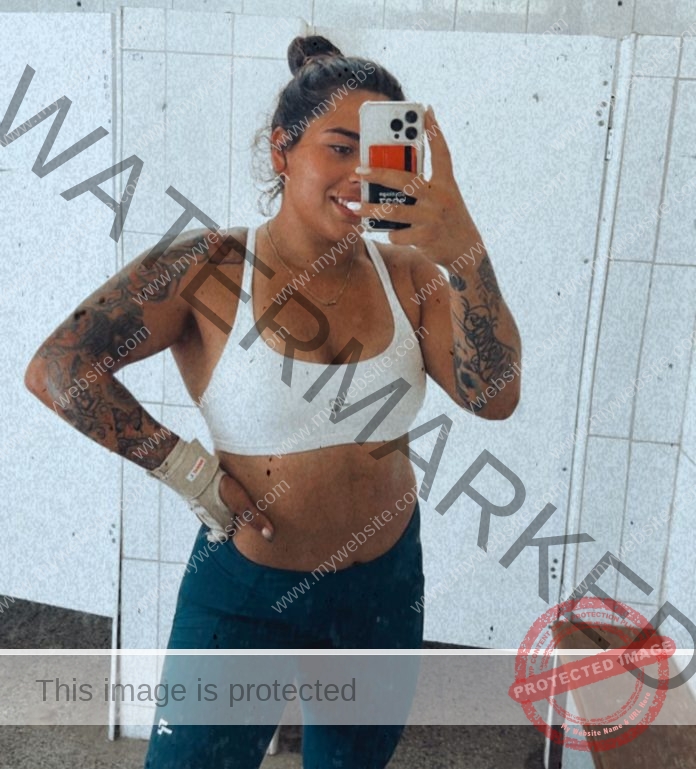 Mariana Walker, hammer thrower from Chile, takes a gym mirror selfie in white sports bra and dark leggings, smiling with hair in a bun.