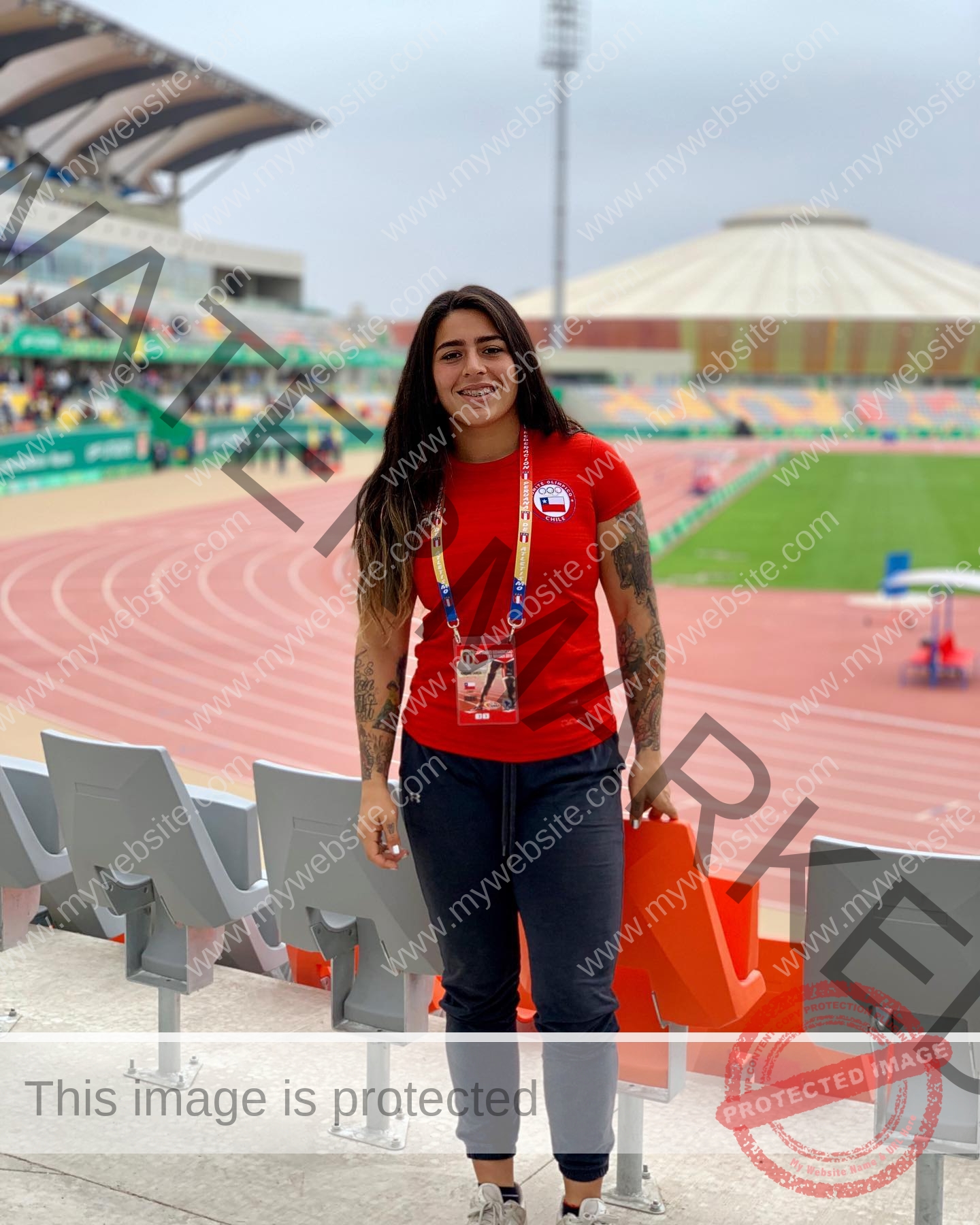 mariana walker-childe-marianawalker_-1914 Mariana Walker, hammer thrower from Chile, stands in a red t-shirt by a stadium track, smiling with long hair and arm tattoos.
