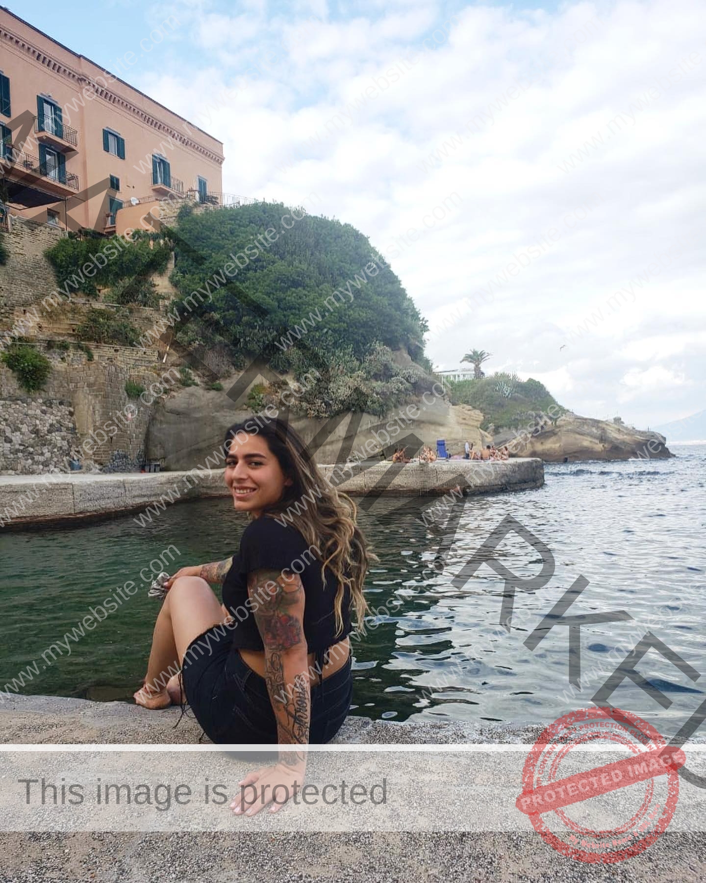 mariana walker-childe-marianawalker_-1938 Mariana Walker, hammer thrower from Chile, smiles with long hair and tattoos on a stone ledge by water, greenery and sky behind.