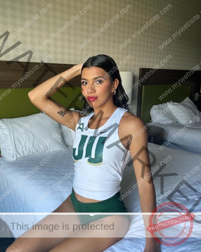 Mariandree Chacon in athletic wear with JU on her top sits confidently on a hotel bed, one arm raised, long dark hair, bold makeup.