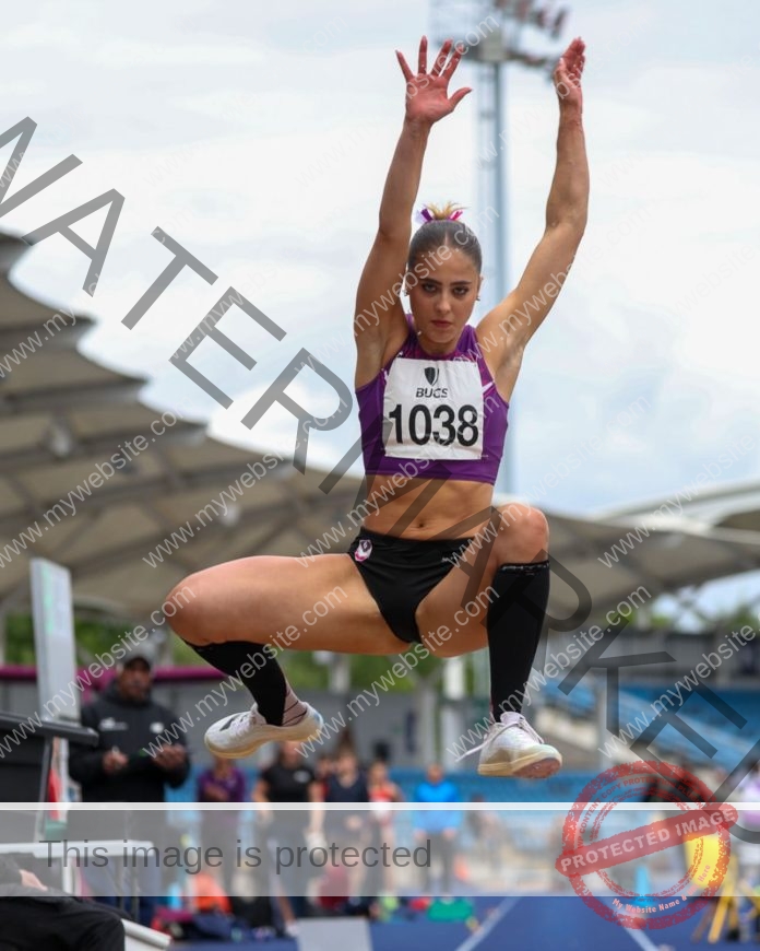 Molly Palmer Molly Palmer, a female athlete from England, soars mid-air in long jump, wearing purple crop top, black shorts, knee-high socks. Stadium and crowd behind.