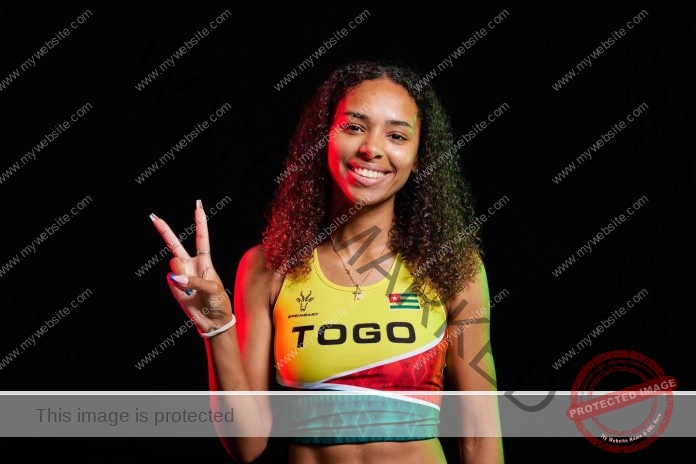 Naomi Akakpo, a smiling female athlete with curly hair, wears a "TOGO" sports top and flashes a peace sign against black backdrop.