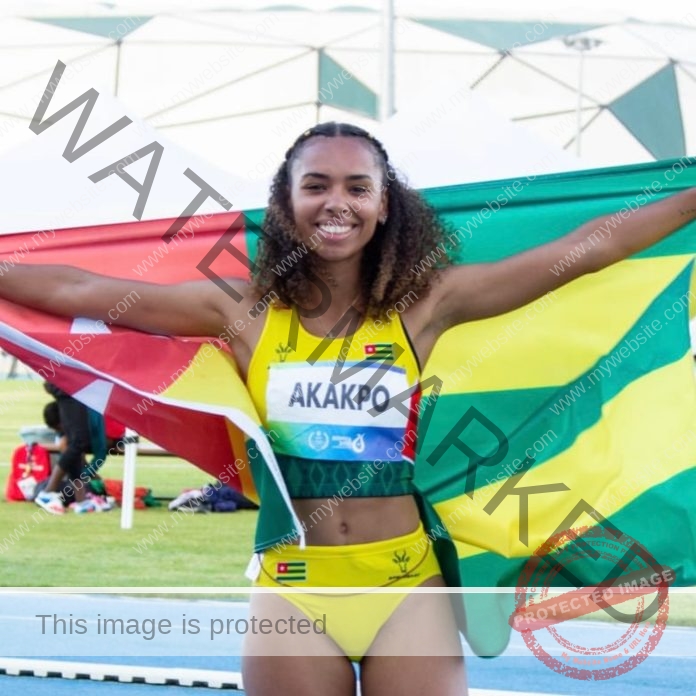 Naomi Akakpo A female track and field athlete in a yellow uniform, labeled Naomi Akakpo, smiles holding a large Togo flag on the track.
