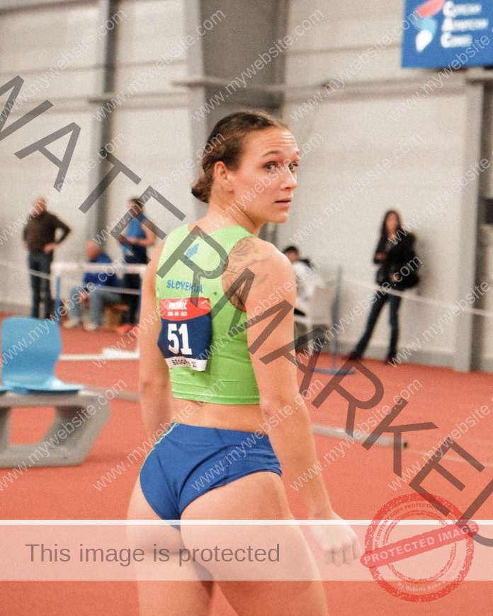 Nika Glojnaric, a female track athlete in a green top and blue shorts, looks over her shoulder on an indoor track with bib 51.