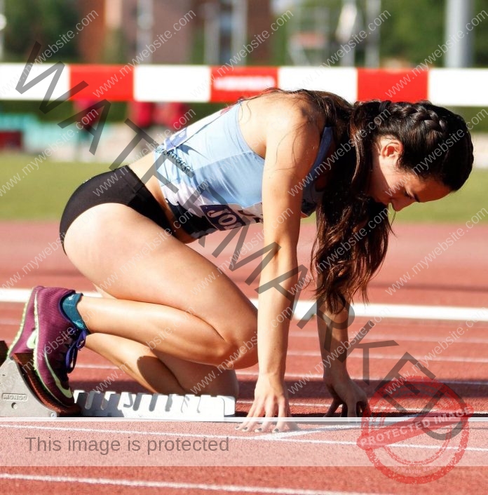 Paula Soria, a female track and field athlete in sportswear, crouches at the starting block on a sunlit Spanish running track.