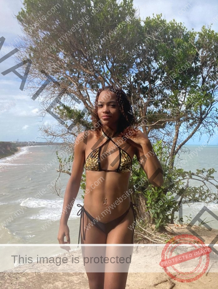 Raylla Vieira of Brazil standing on the ebach in a floral print bikini, trees in the background, playing with her hair.