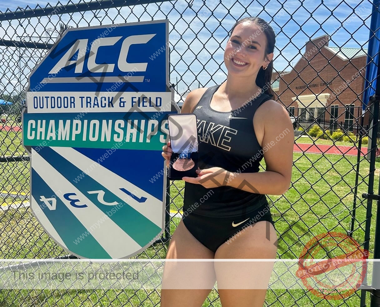 rosa-gergely-wake-forest-th-ro_gerg-08329 Rosa Gergely A female athlete in a black WAKE Forest uniform smiles, holding a medal by an ACC Outdoor Track sign on a sunny day.