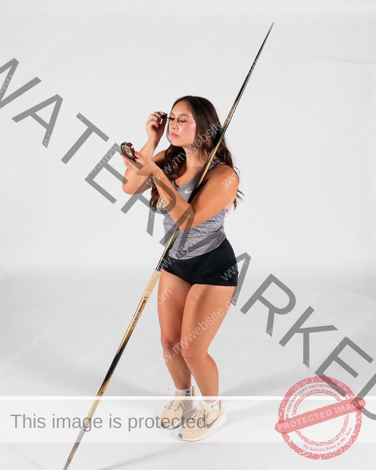 rosa-gergely-wake-forest-th-ro_gerg-1993 Rosa Gergely, javelin thrower from Wake Forest, holds a javelin upright while applying makeup with a compact mirror on white background.