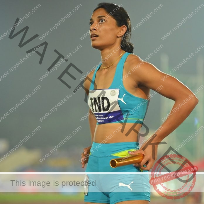 Sakshi Chavan Track and field athlete Sakshi Chavan stands on the track in a blue India uniform, holding a relay baton, focused.