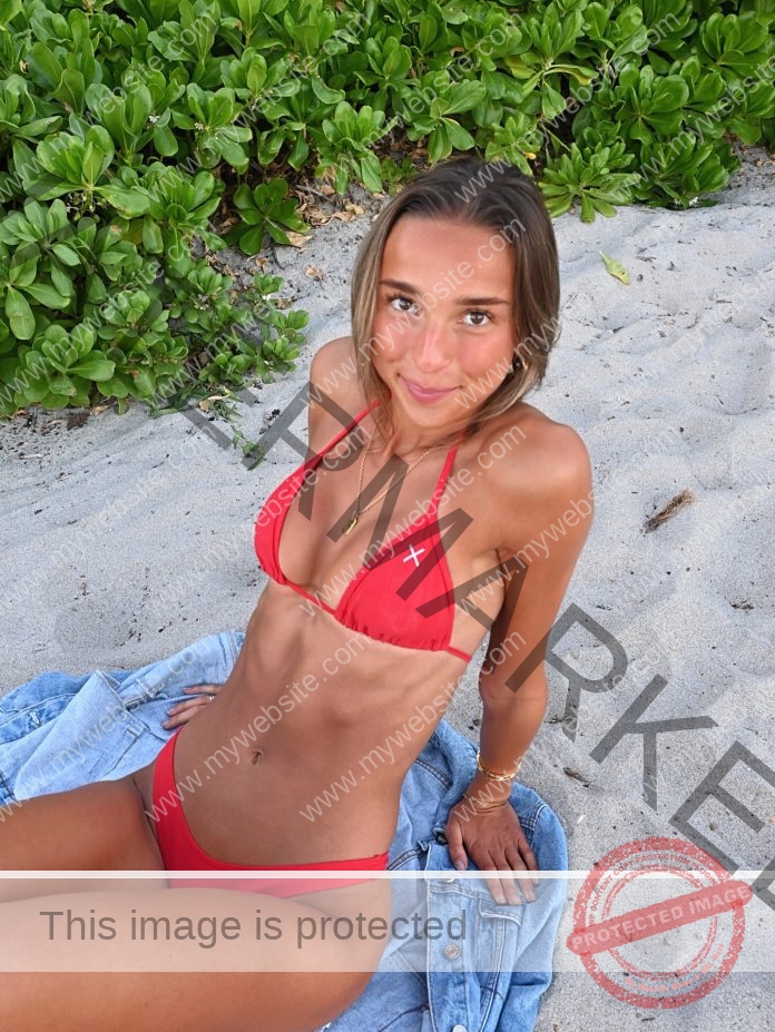 Sammi Jo Payne in a red bikini sits on sandy beach, denim jacket beneath her, smiles at camera—soccer player vibes on a sunny day.