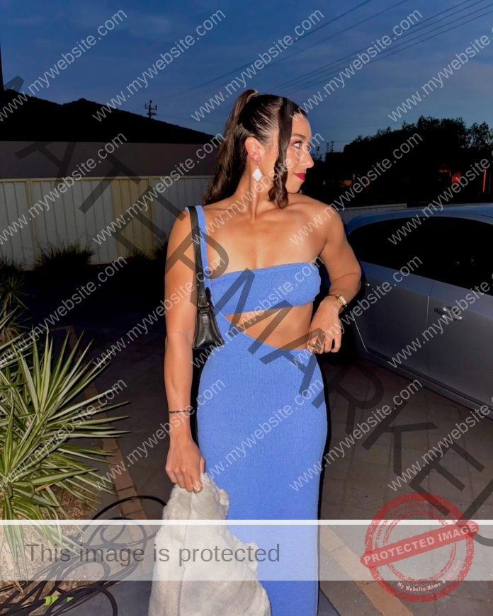 Sarah Schwarz Sarah Schwarz at dusk in Australia, wearing a blue one-shoulder cutout dress, holding gray garment and black bag by a car.