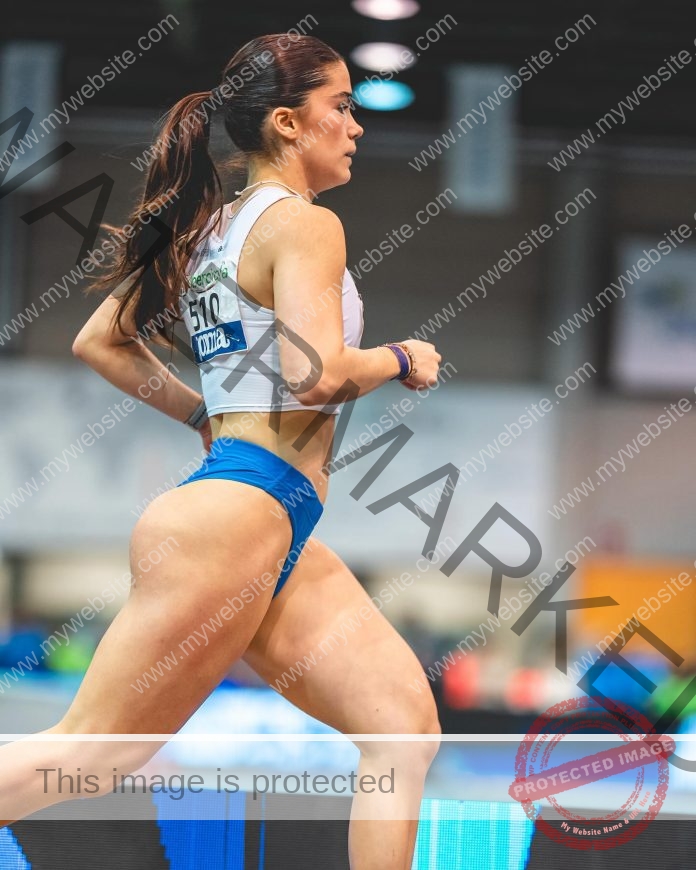 Sofia Cosculluela Sofia Cosculluela, Spanish track athlete, runs indoors in blue shorts and a white crop top, focused mid-stride.