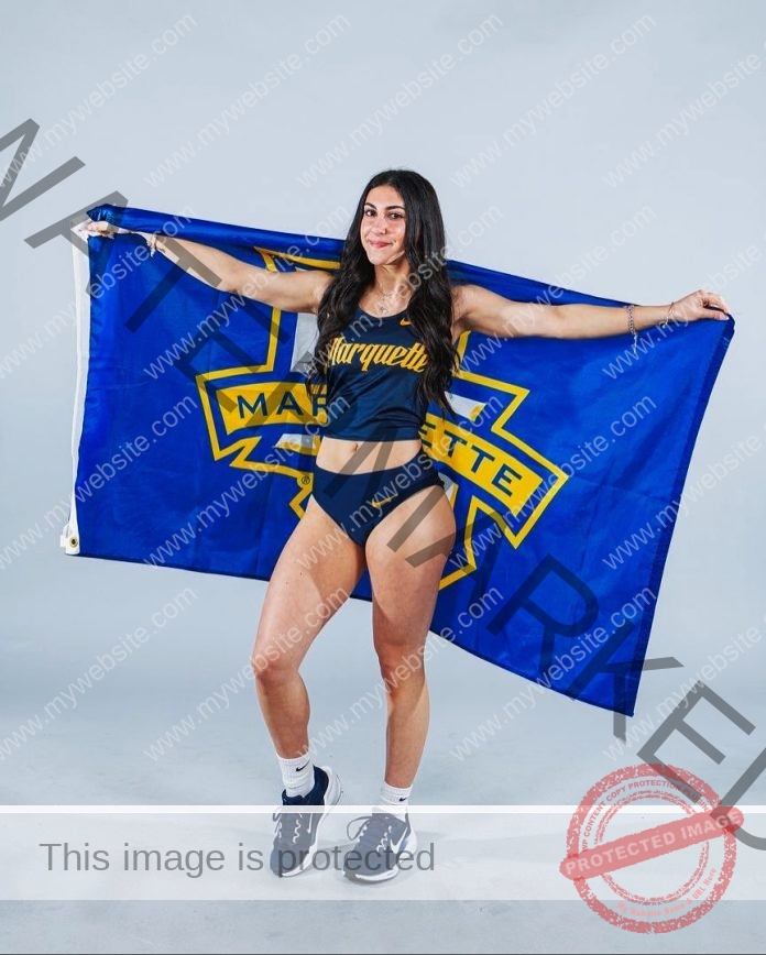 Sophia Abraham standing in front of a white background in her track uniform, holding her school flag up behind her.