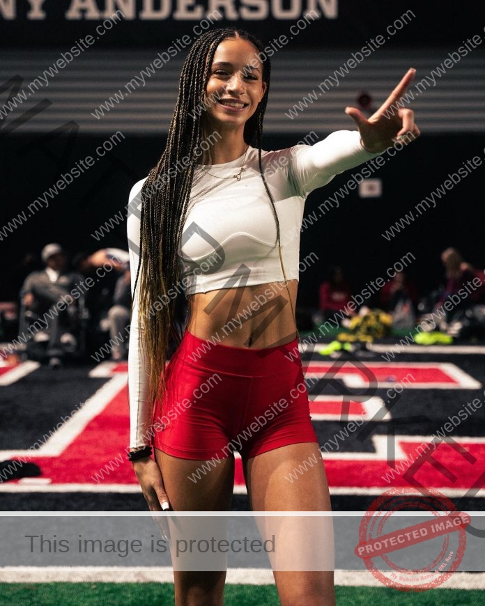 Tamiah Washington, a smiling woman with long braids, white crop top, and red shorts, stands flashing a peace sign on an indoor athletic field.