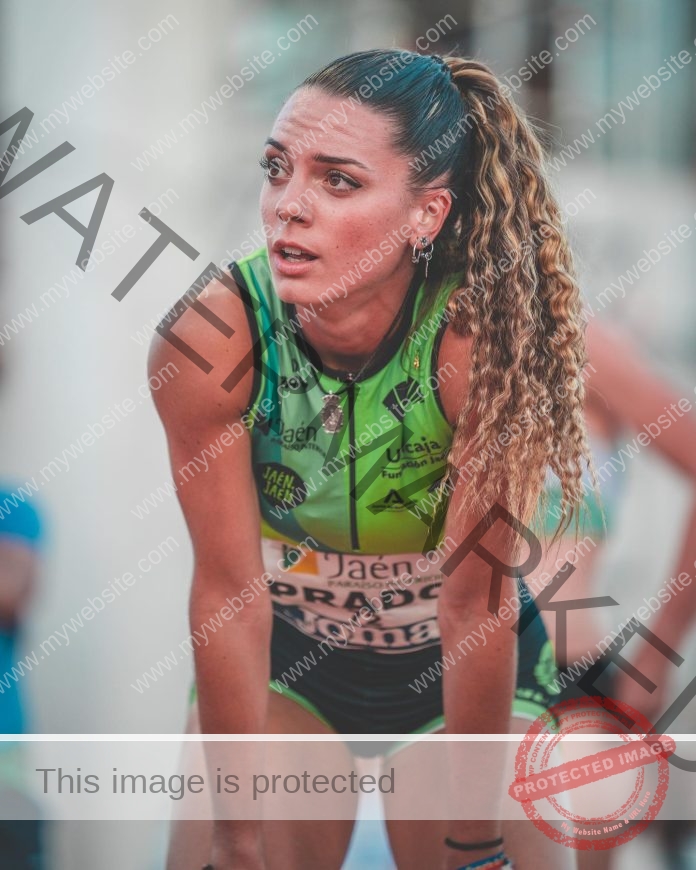 Valme Prado, a female track and field athlete from Spain in a green and black uniform, leans forward catching her breath outdoors.