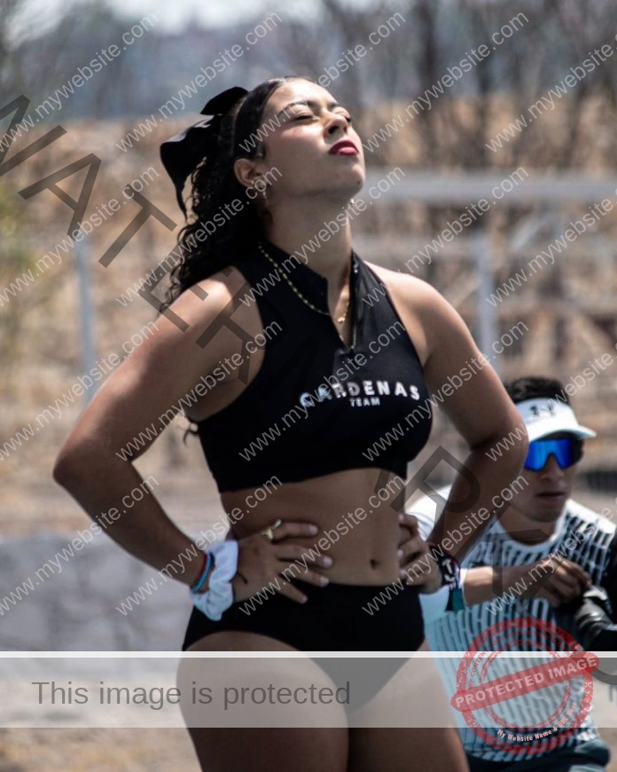 Vianey Santos, a woman in black sportswear stands hands on hips, eyes closed. Her top reads CARDENAS TEAM for Mexico; man in background.