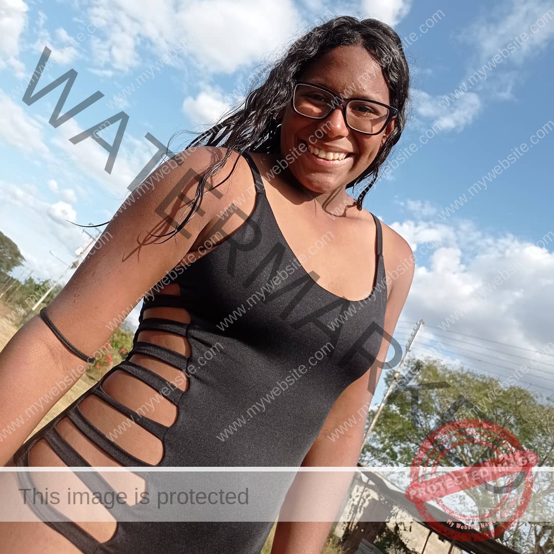 yenni-veroes-venezuela-yenni__throw-002 Alejandra Veroes, a person with long dark hair and glasses, smiles outdoors in a black swimsuit with side cutouts. Sky and greenery behind.