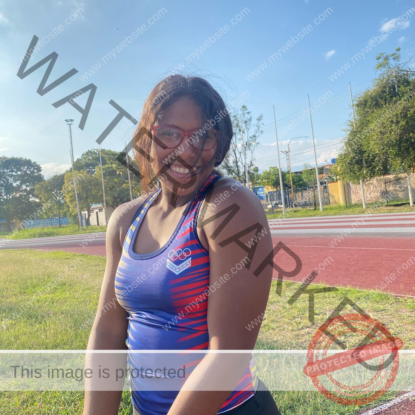 yenni-veroes-venezuela-yenni__throw-016 Alejandra Veroes, smiling in athletic gear, stands on a sunny track in glasses and a blue Olympic tank top; trees and fence behind.