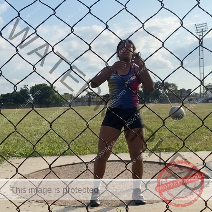 yenni-veroes-venezuela-yenni__throw-017 Yenni Veroes, a female athlete, stands in a throwing circle ready to spin and throw a hammer on a grassy field under blue sky.