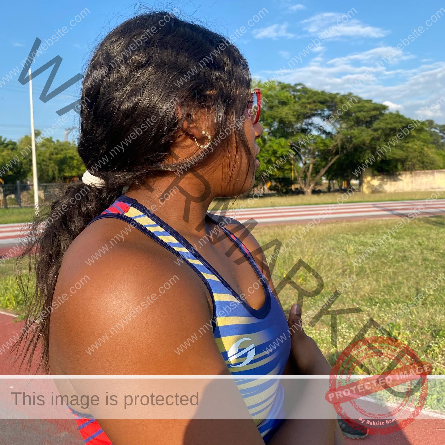 yenni-veroes-venezuela-yenni__throw-018 Alejandra Veroes, a woman with dark hair in a ponytail and blue and yellow tank top, stands on a track, arms crossed, outdoors.