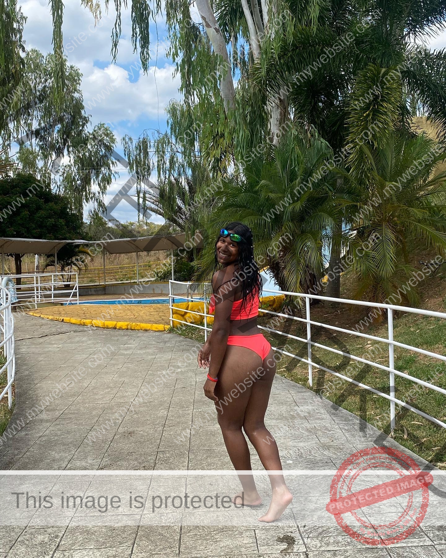 yenni-veroes-venezuela-yenni__throw-032 Alejandra Veroes A woman in a red bikini stands on a paved path near a pool, smiling with sunglasses on her head and palm trees behind.