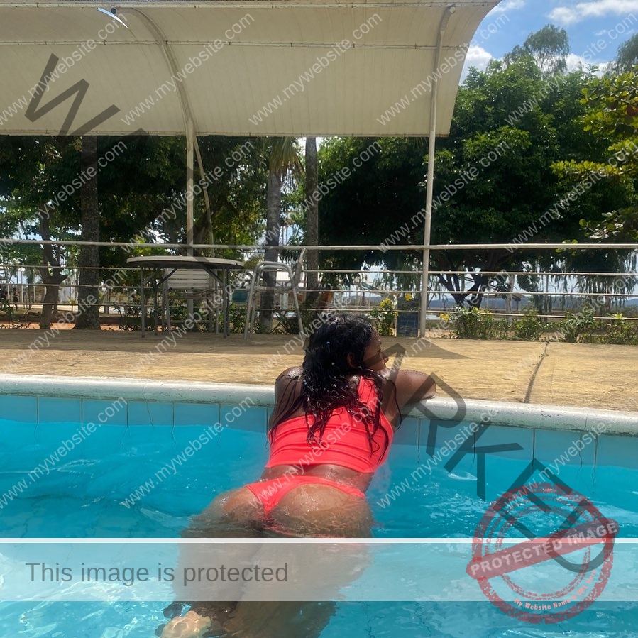 yenni-veroes-venezuela-yenni__throw-035 Alejandra Veroes A woman in a red swimsuit leans on the pool edge, looking away, with trees and outdoor seating under bright daylight.