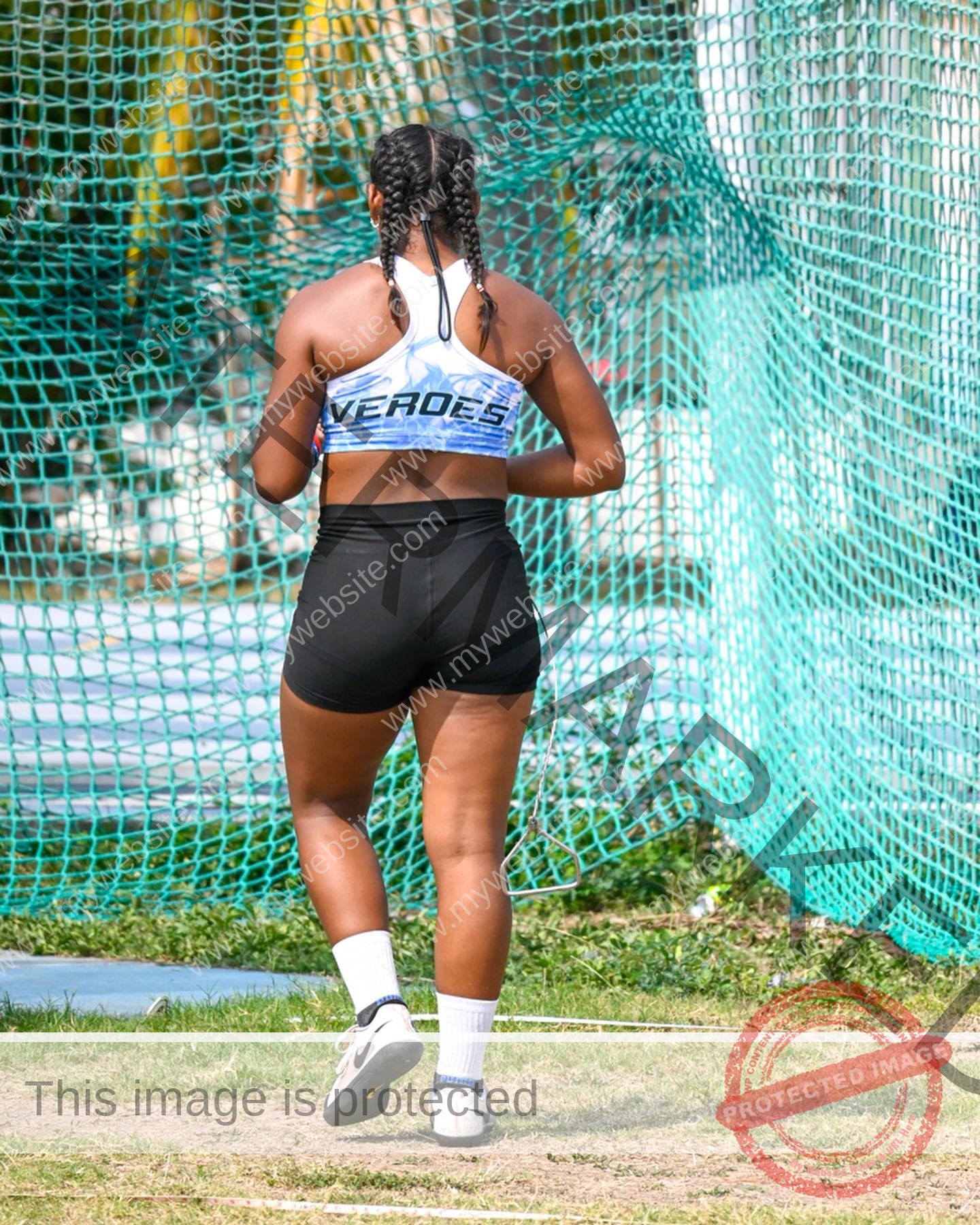 yenni-veroes-venezuela-yenni__throw-037 Alejandra Veroes stands on grass facing away, in a blue sports bra, black shorts, white socks, with a net and palm trees behind.