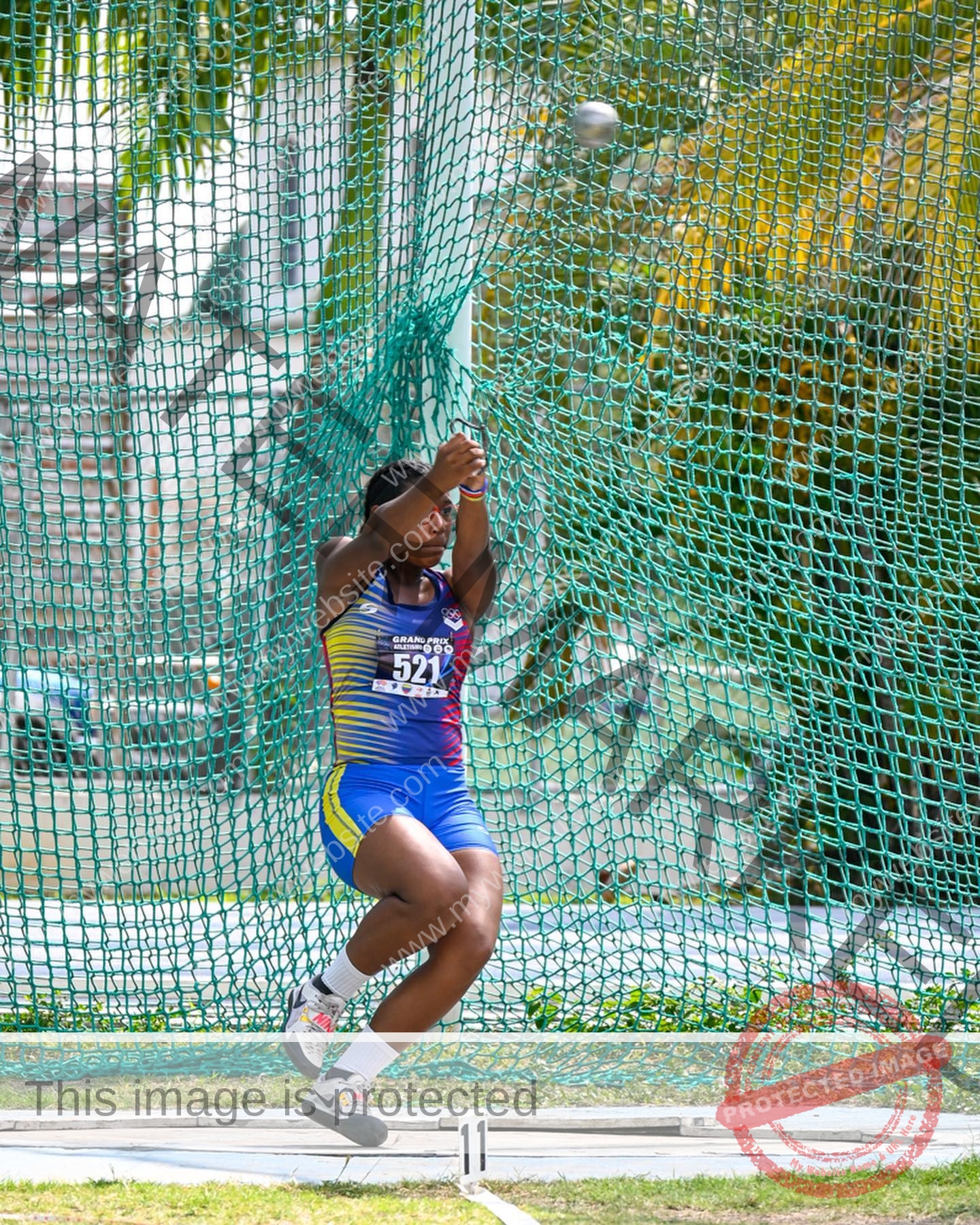 yenni-veroes-venezuela-yenni__throw-038 Yenni Veroes A female athlete in a blue and yellow uniform swings a hammer during a track and field event, with green netting and palm trees behind.
