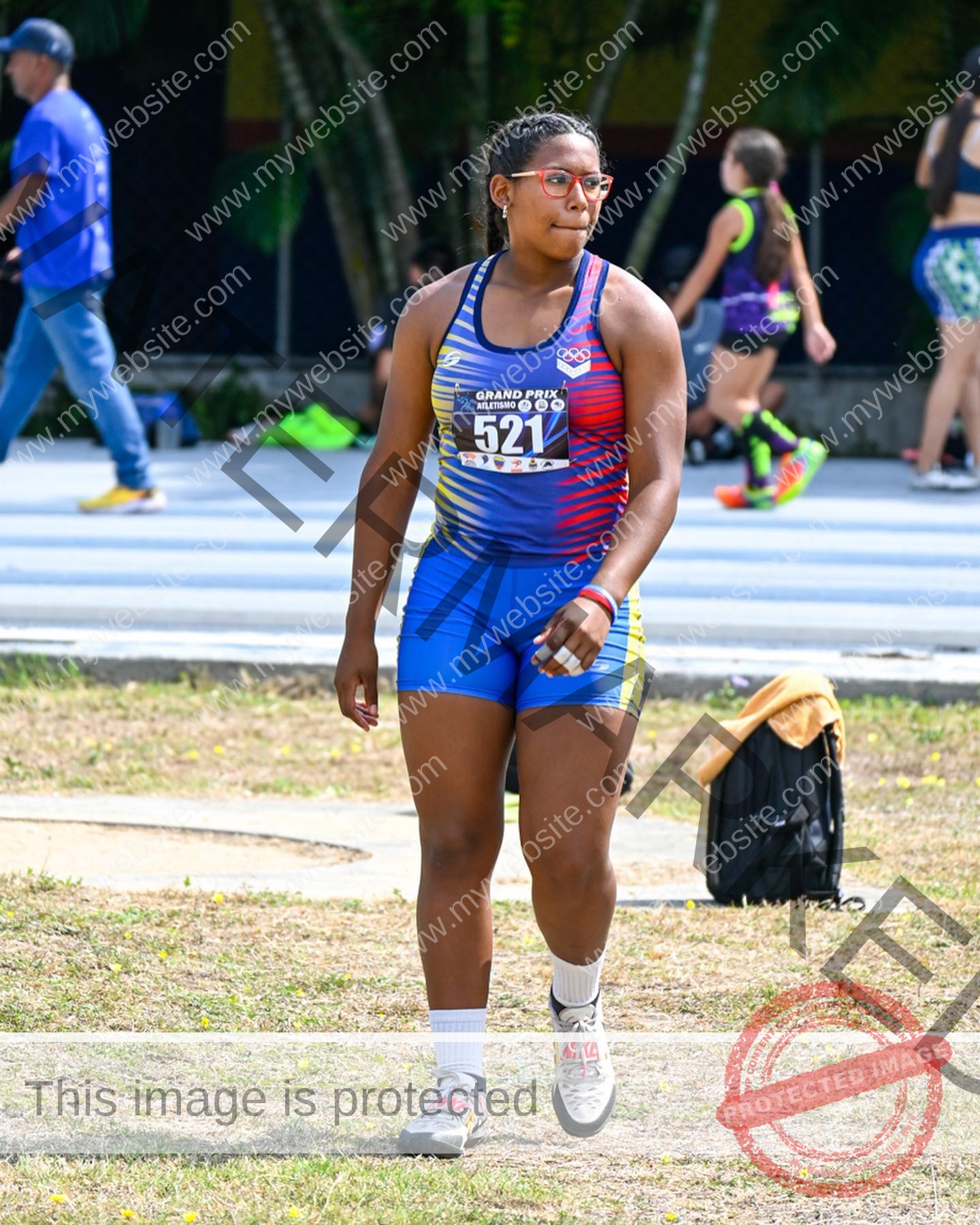 yenni-veroes-venezuela-yenni__throw-046 Yenni Veroes A female athlete in a colorful track and field uniform and red glasses walks on the grass at an outdoor sports event, with other athletes and people visible in the background.