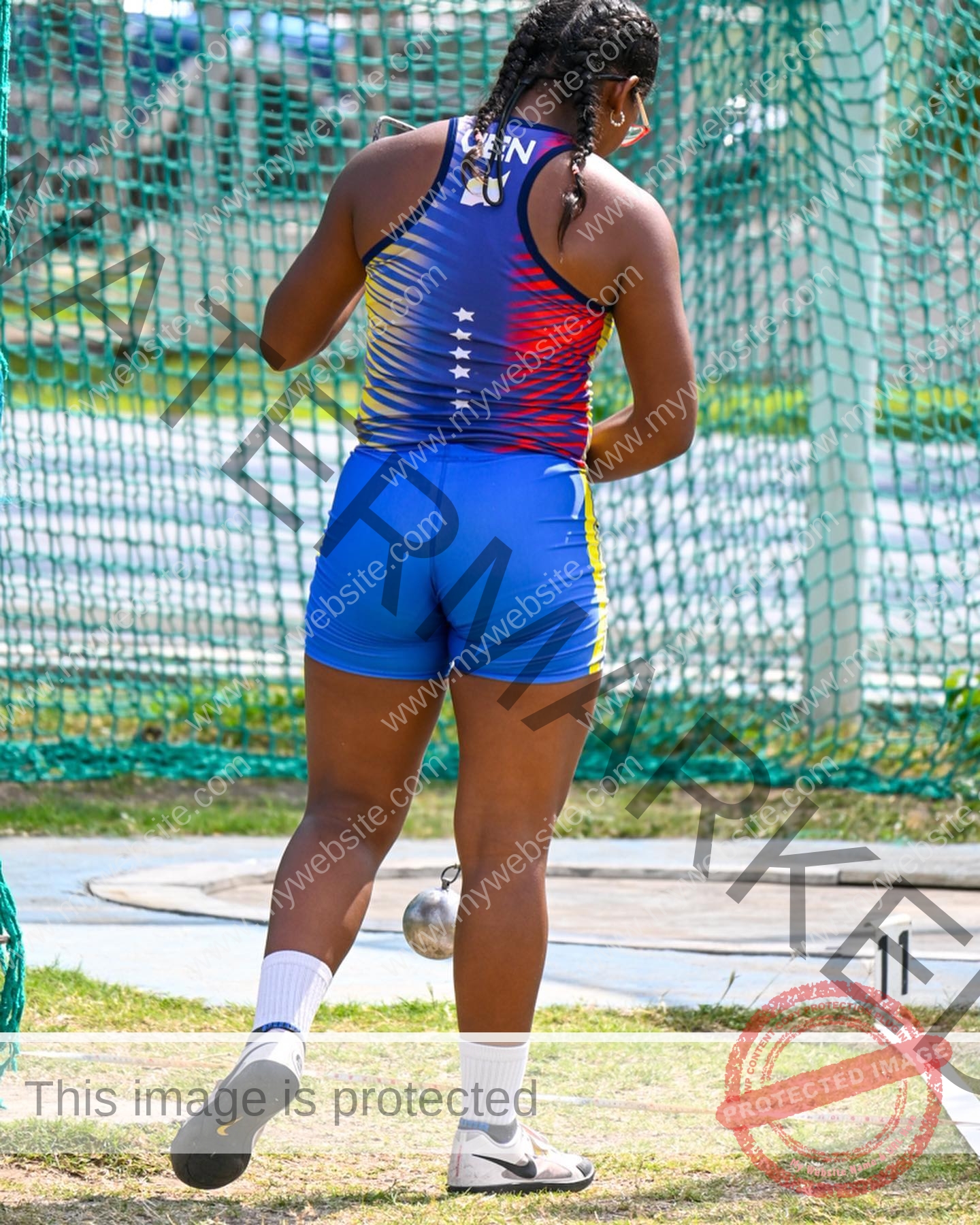 yenni-veroes-venezuela-yenni__throw-048 Yenni Veroes, a female athlete in blue, red, and yellow, stands on a throwing circle with a hammer, facing away near the safety net.