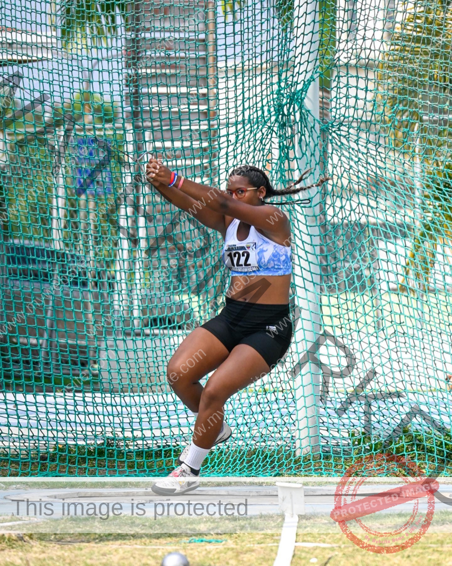yenni-veroes-venezuela-yenni__throw-050 Yenni Veroes, a female athlete in a blue and white sports bra and black shorts, prepares to throw a discus in a netted cage.