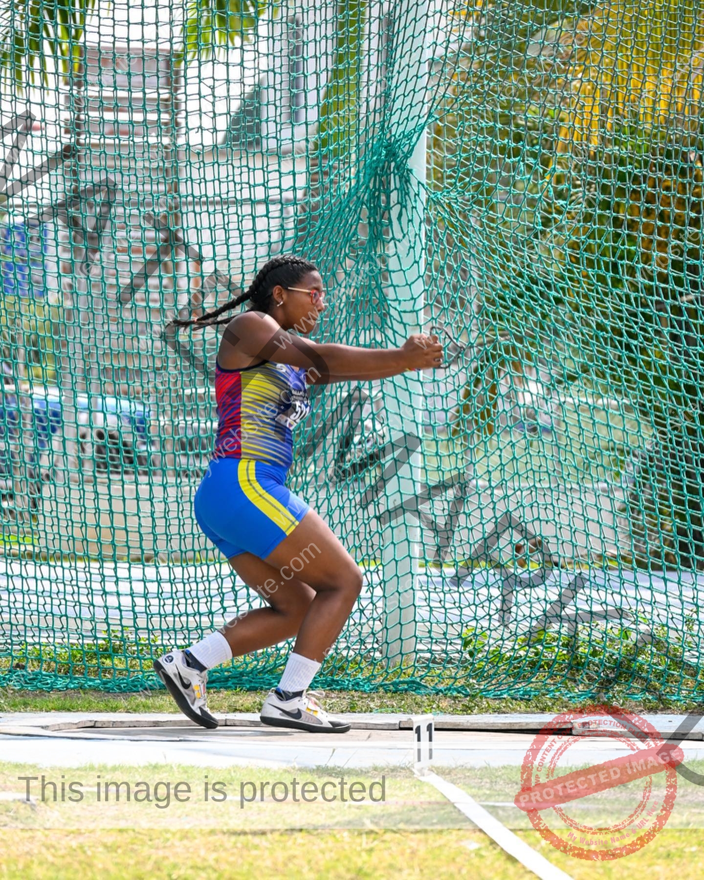 yenni-veroes-venezuela-yenni__throw-053 Alejandra Veroes, an athlete in a colorful uniform, prepares to throw a discus in a netted cage at an outdoor track and field event.
