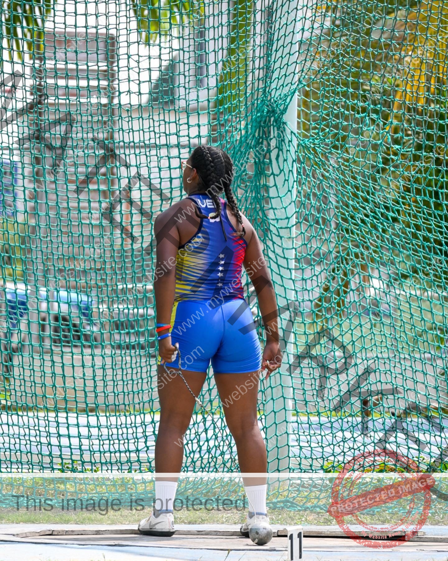 yenni-veroes-venezuela-yenni__throw-054 Alejandra Veroes A female athlete in blue and multicolored gear stands back to camera, holding a hammer throw chain by the green net.