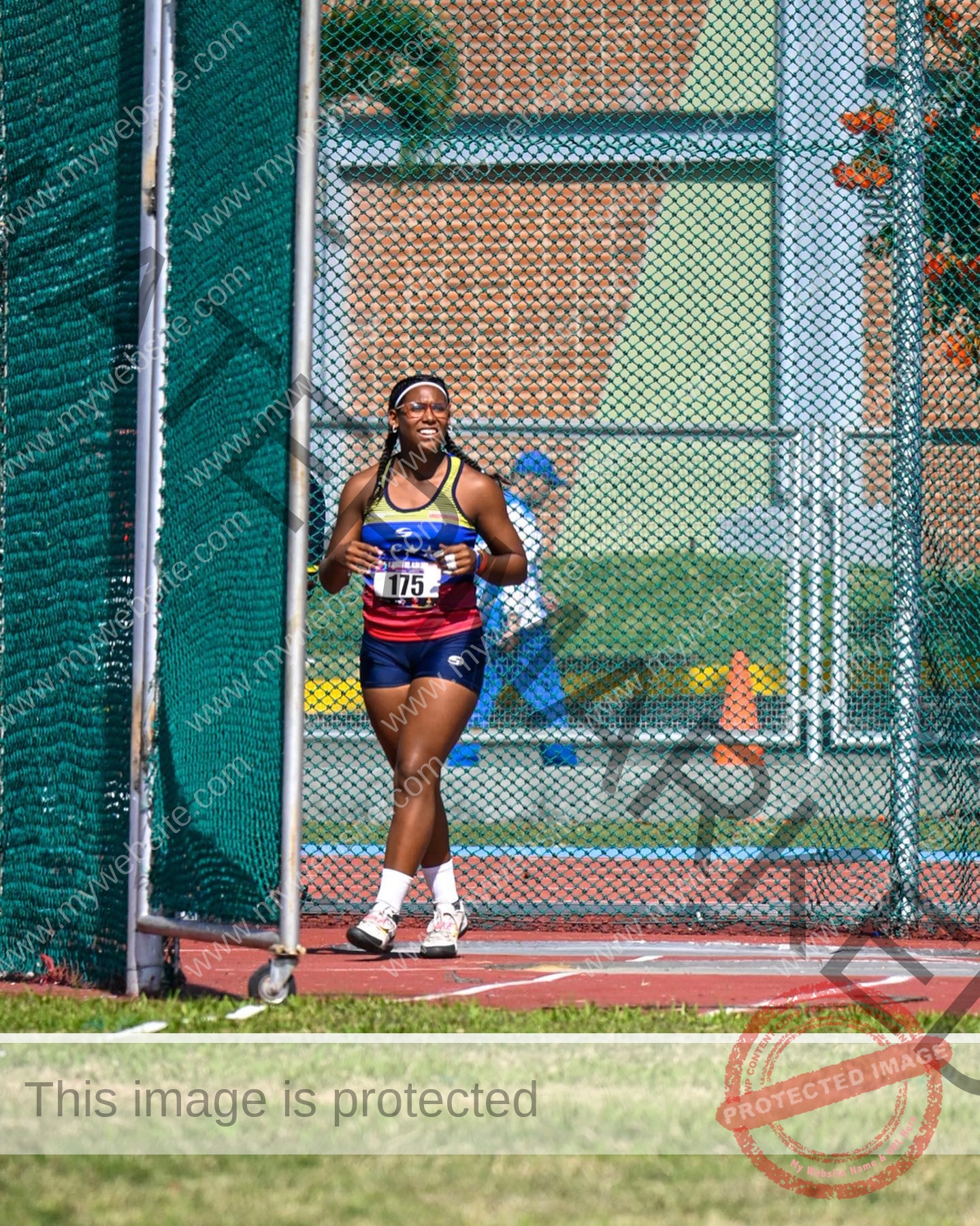 yenni-veroes-venezuela-yenni__throw-064 Alejandra Veroes A female athlete in a blue and red uniform, number 175, smiles walking on a track near a green net and chain-link fence.