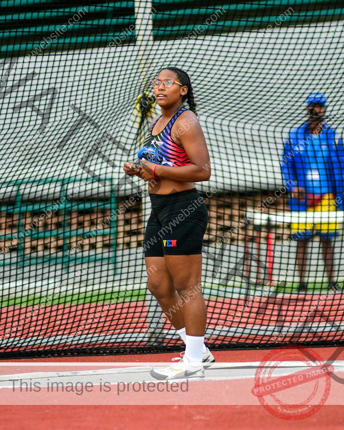 yenni-veroes-venezuela-yenni__throw-079 Alejandra Veroes, a female athlete in colorful sportswear and glasses, stands on a track ready to compete; a net is behind her.