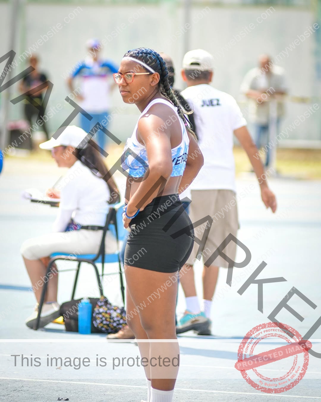 yenni-veroes-venezuela-yenni__throw-087 Alejandra Veroes, a young athlete in sportswear, stands on a track with hands on hips, braided hair, glasses; blurred crowd behind.