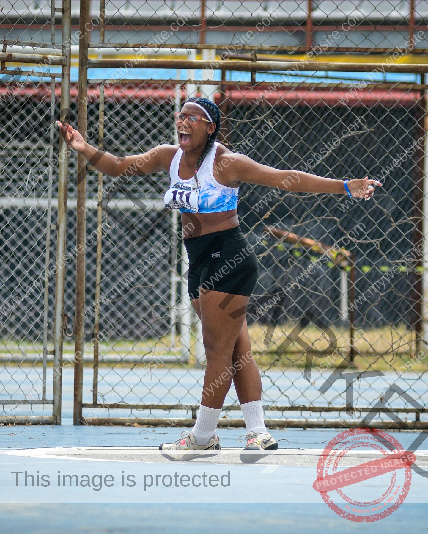 yenni-veroes-venezuela-yenni__throw-088 Yenni Veroes A woman wearing athletic clothes and sneakers stands on a sports field, arms outstretched and smiling, with buildings behind her.