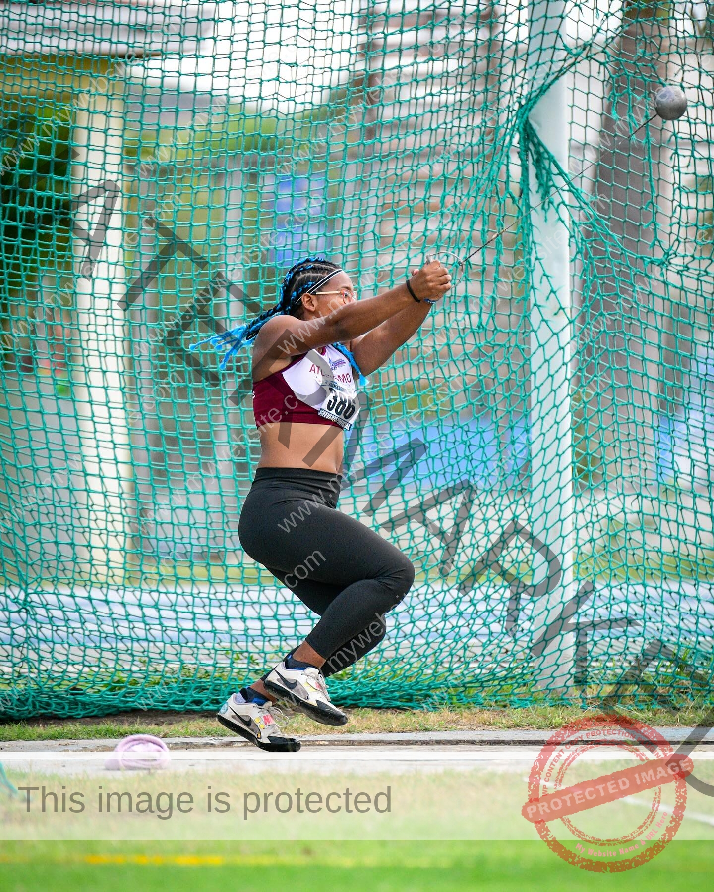 yenni-veroes-venezuela-yenni__throw-090 Yenni Veroes, a female athlete in athletic wear, swings a hammer in a caged area at a track meet, mid-motion and focused.