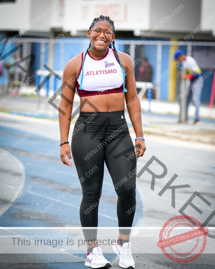 Yenni Veroes A smiling athlete wearing a crop top labeled ATLETISMO, black leggings, and sneakers stands on a running track, with buildings and another person visible in the blurred background.