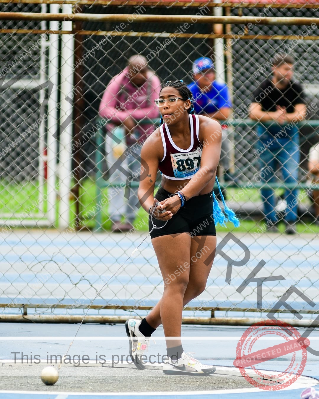 yenni-veroes-venezuela-yenni__throw-108 Alejandra Veroes, a female athlete in sportswear and a bib, readies to throw a hammer at a track event with onlookers behind.