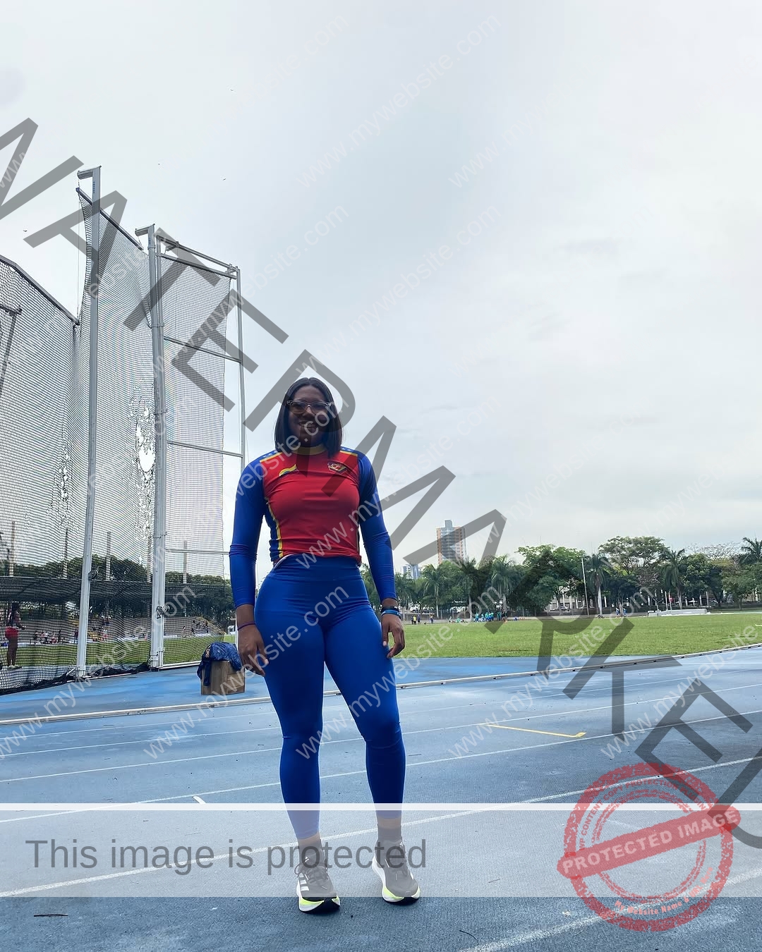 yenni-veroes-venezuela-yenni__throw-117 Yenni Veroes A person wearing a red and blue athletic outfit stands smiling on an outdoor running track, with sports nets and trees behind.