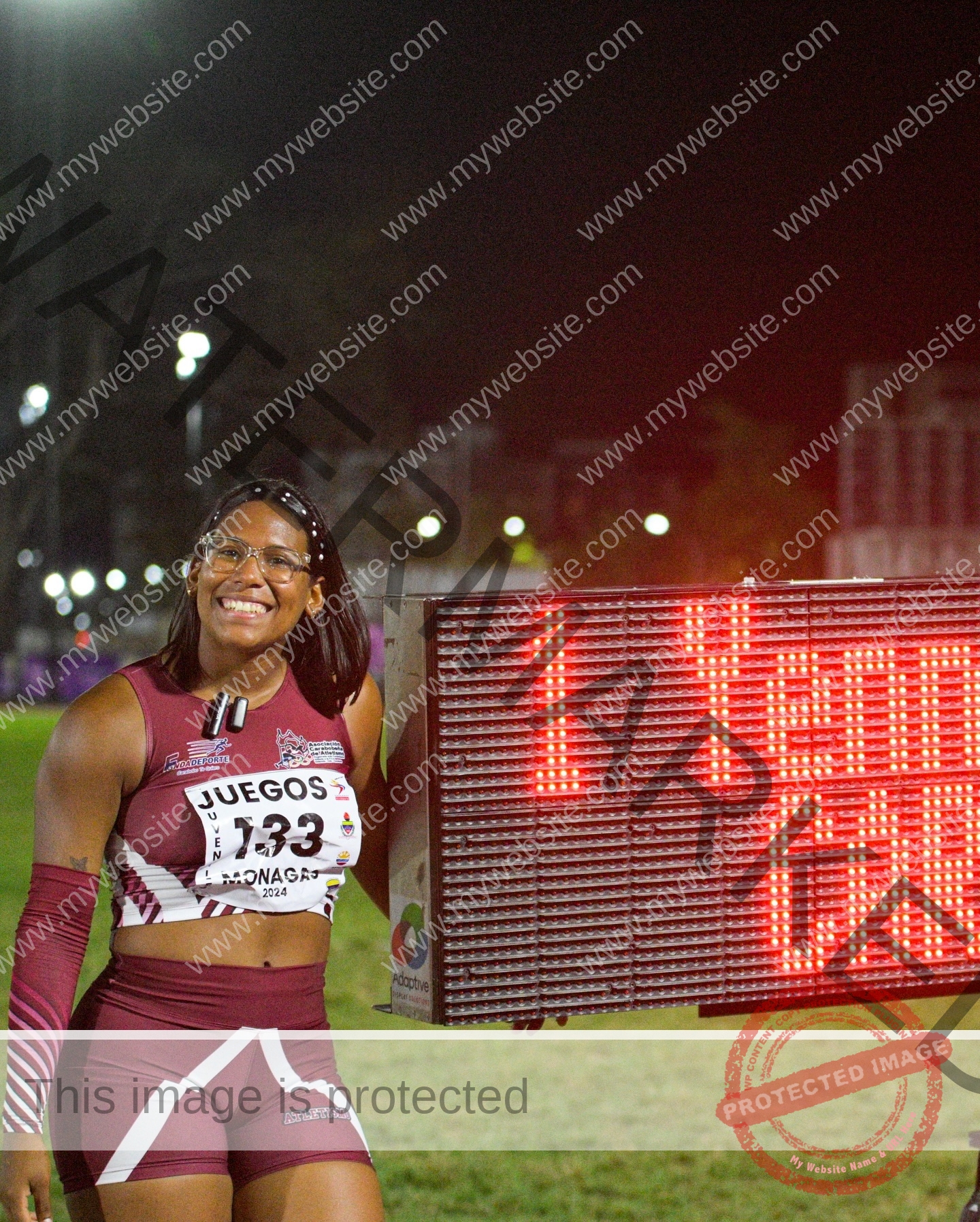 yenni-veroes-venezuela-yenni__throw-128 Yenni Veroes, a smiling female athlete in maroon and white track uniform, stands by a lit electronic scoreboard at night.
