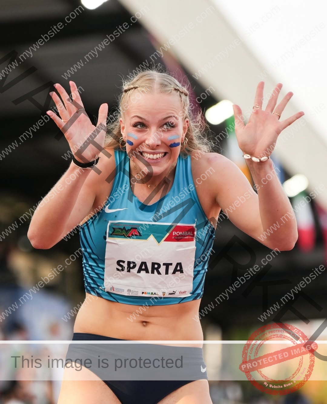 amaya-kruse-jorgensen-denmark-amayajorgensen-0260 Amaya Kruse Jorgensen, track and field star from Denmark, in a blue crop top and navy shorts, smiles and raises both hands.