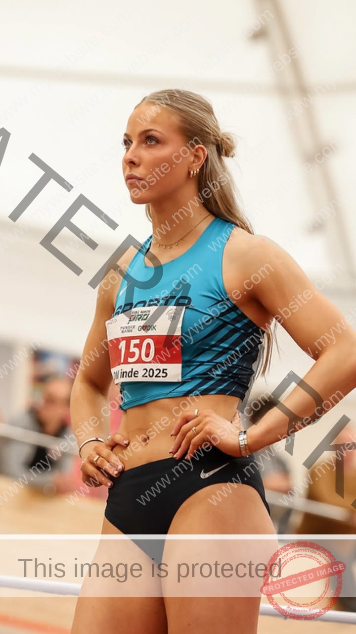 amaya-kruse-jorgensen-denmark-amayajorgensen-0287 Amaya Kruse Jorgensen, track and field star from Denmark, stands focused in blue crop top, bib 150/Winde 2025 at indoor event.