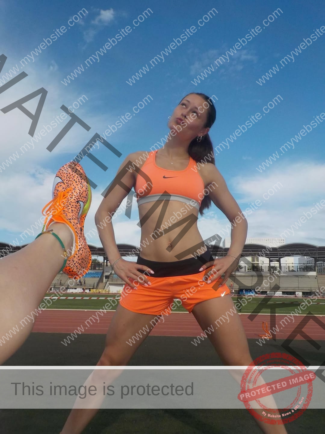amaya-kruse-jorgensen-denmark-amayajorgensen-0306 Amaya Kruse Jorgensen, track and field star from Denmark, poses on a track in orange sportswear; a shoe is raised in the foreground.