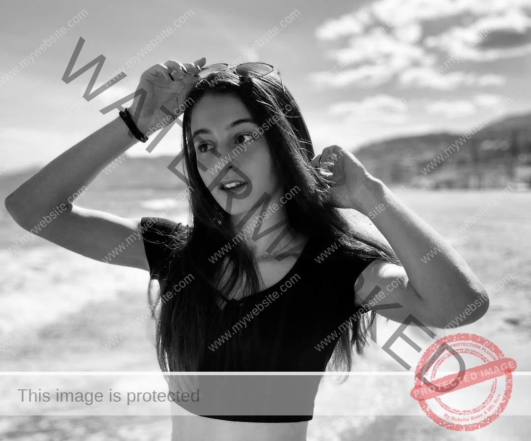 anabel-vitale-italy-anabelvitale-0384 Anabel Vitale, track and field star from Italy, stands on a beach in a black crop top, holding sunglasses. Waves and clouds behind.