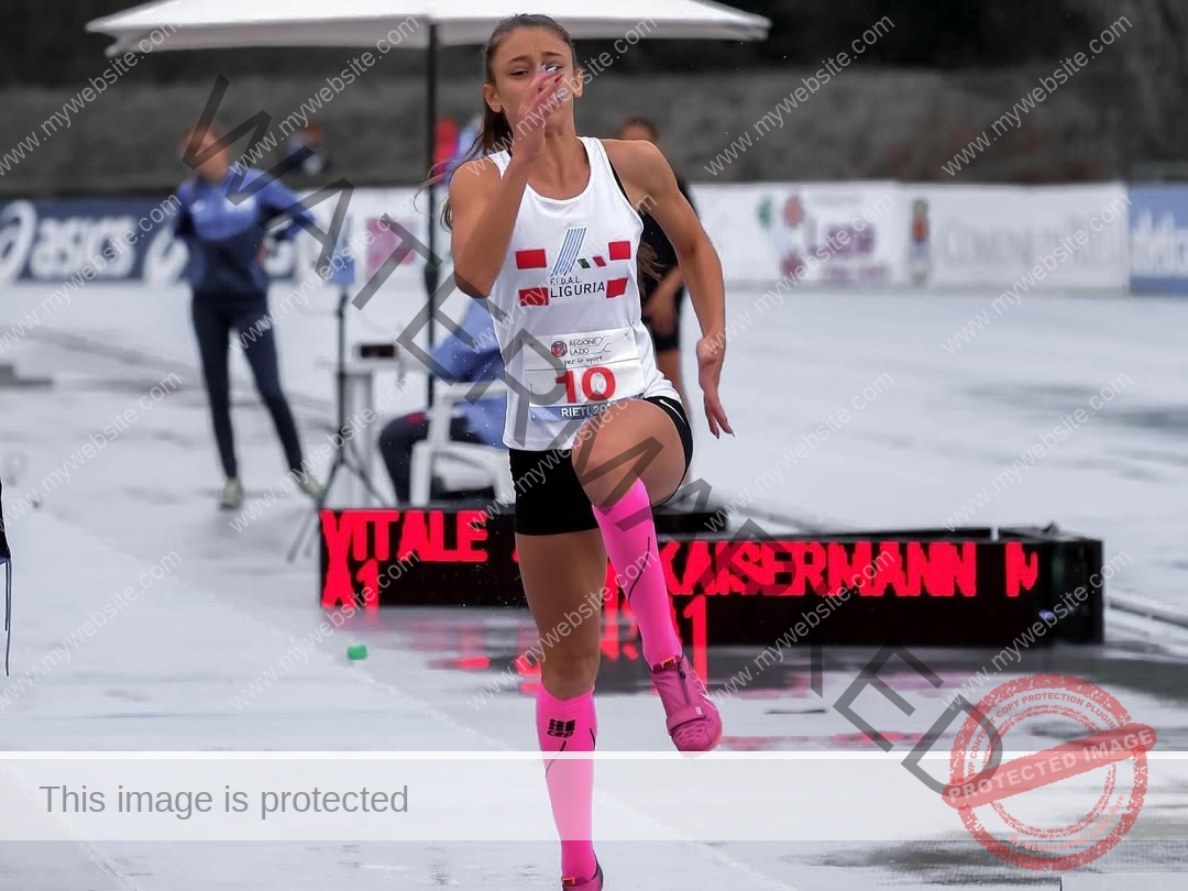 anabel-vitale-italy-anabelvitale-0390 Anabel Vitale, track and field star from Italy, in a white jersey and pink socks jumps on a wet track; others and umbrella behind.